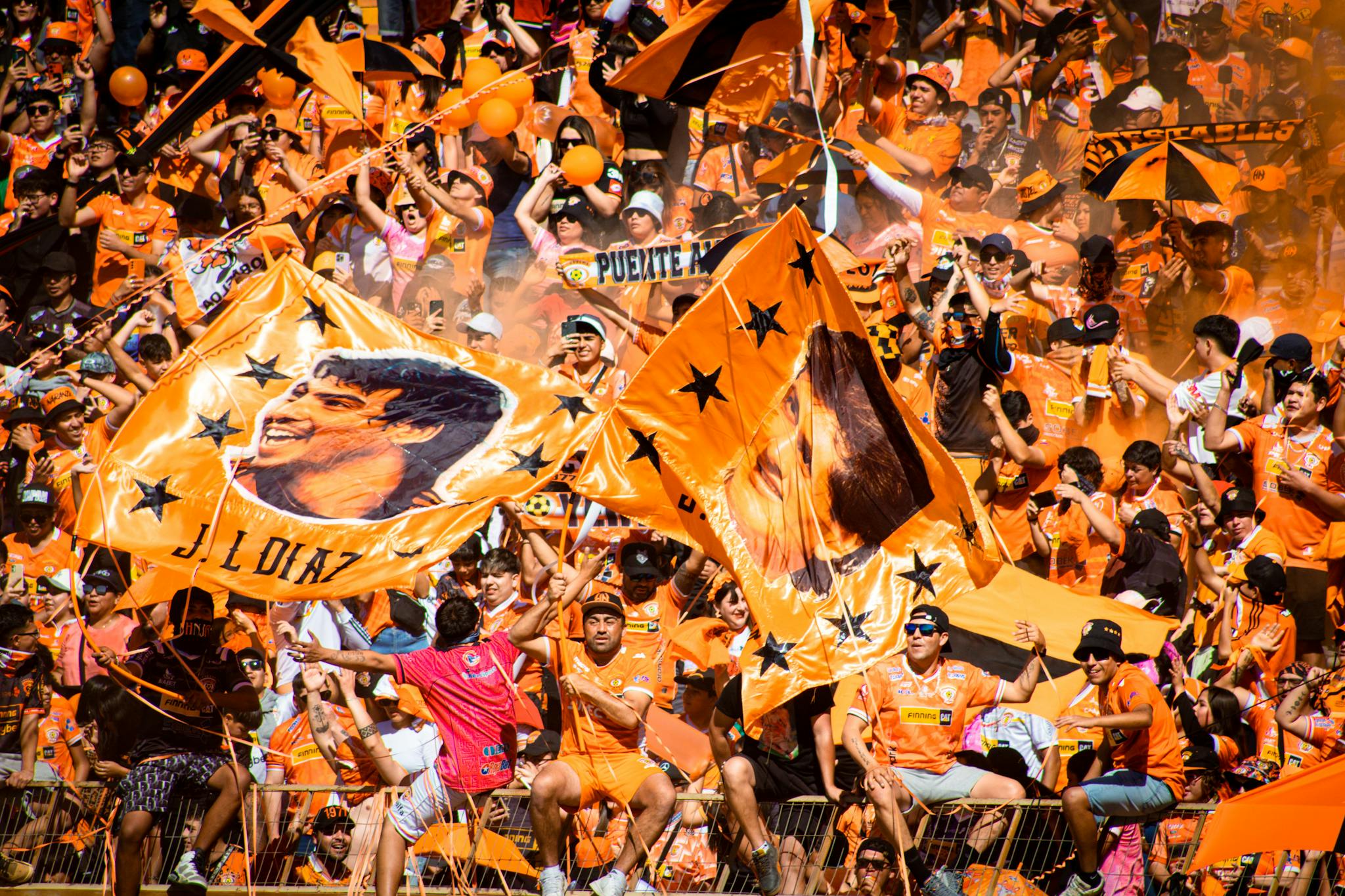 Energetic football fans in Calama, Chile, cheering with flags and vibrant orange attire.